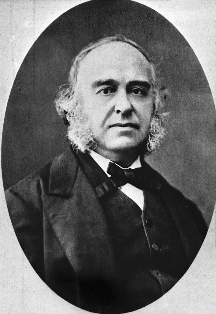 A black and white photo of a man with long hair and mutton chops wearing a black short bowtie and suit and vest.