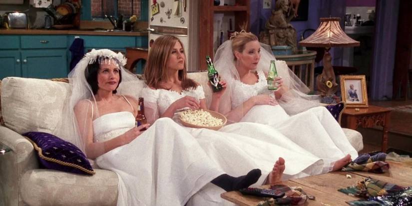 Monica Rachel and Phoebe sitting on the couch in wedding dresses and eating popcorn in Friends