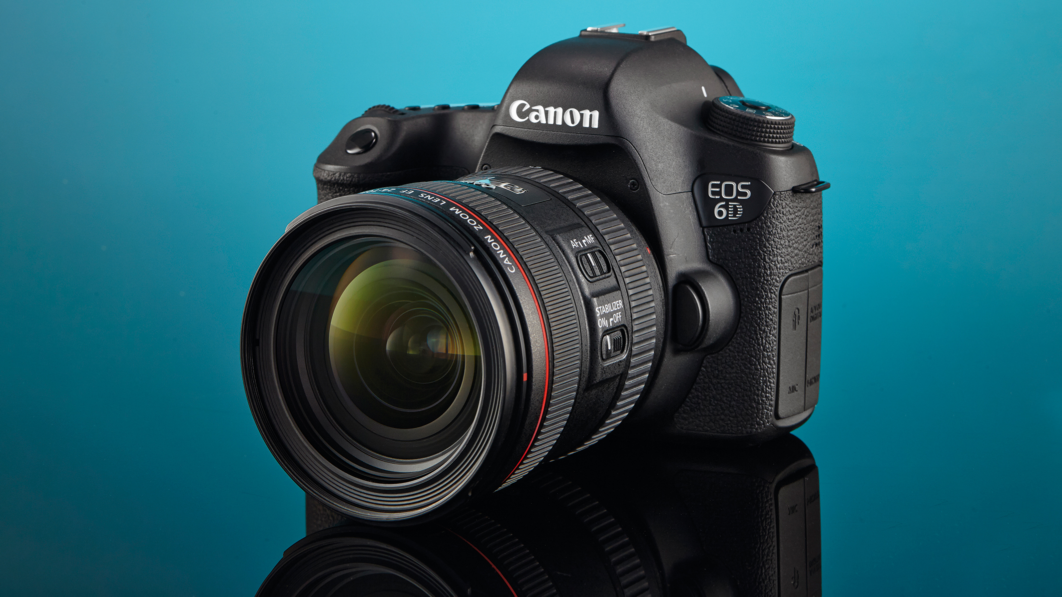 A Canon EOS 6D DSLR camera with a zoom lens, displayed on a reflective surface against a teal background