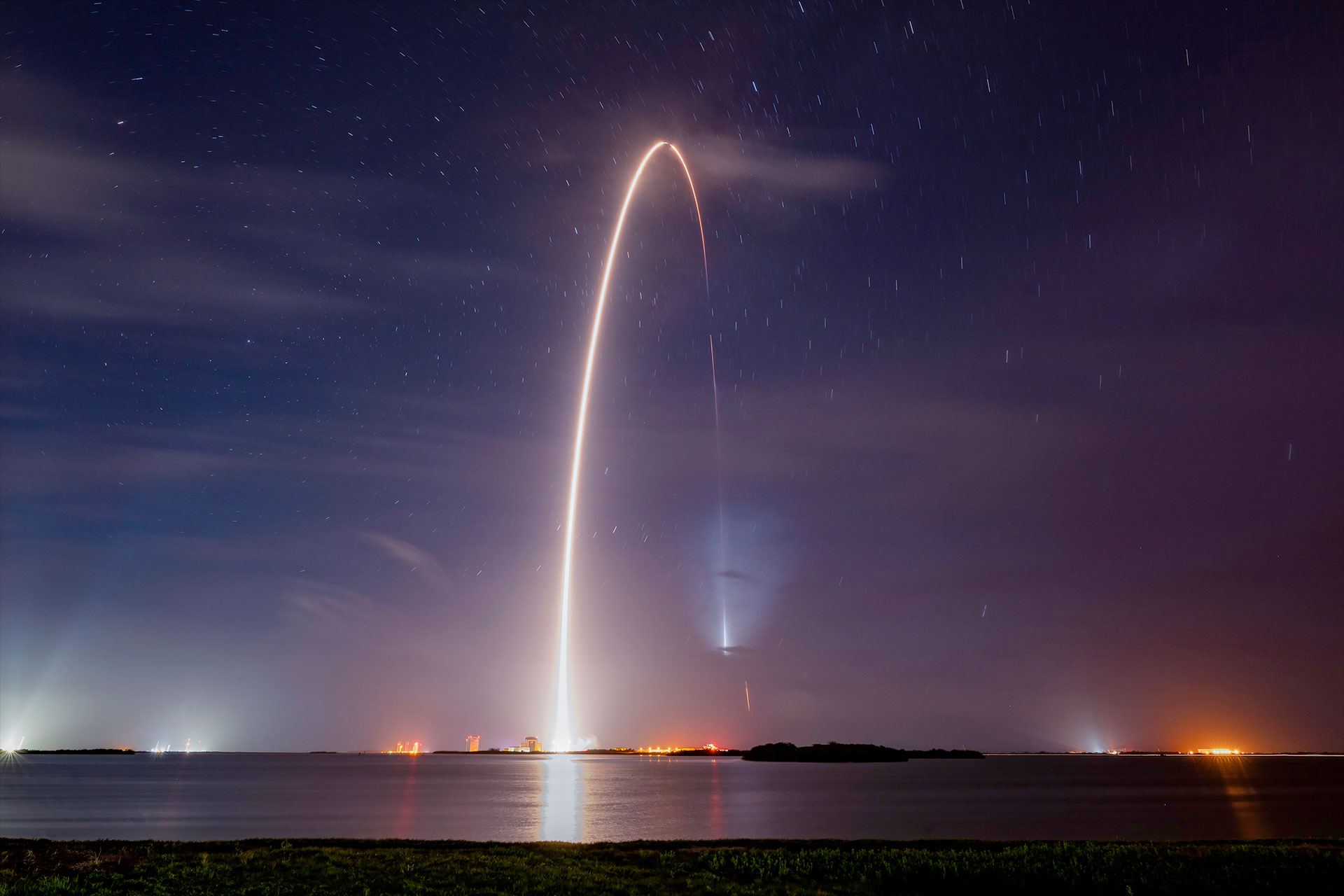 a time lapse exposure captures the glowing arc from the launch and landing of a rocket over a body of water and pre-sunrise purple hazy sky.