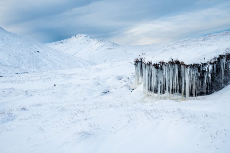 A snow-covered landscape with icy mountains in the background. In the foreground, a structure’s roof is lined with long, hanging icicles, contrasting with the white snow and cloudy sky.