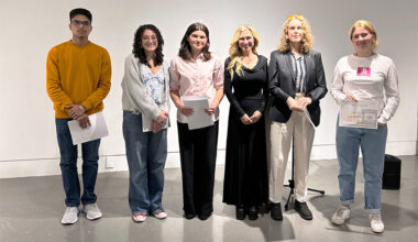 A group of six people stand in a line against a blank wall.
