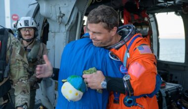 a man in an orange flight suit smiles while carrying a small stuffed moon