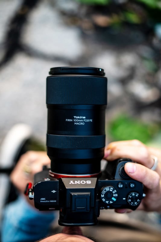 A person holding a Sony camera with a Tokina FIRIN 100mm F2.8 FE macro lens, seen from above with a blurred outdoor background.