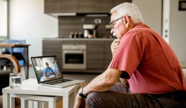 An elderly man in a red shirt attentively watches a video call with V A TelePulmonology on a laptop in a modern kitchen. A glass of water sits on a white table near him.