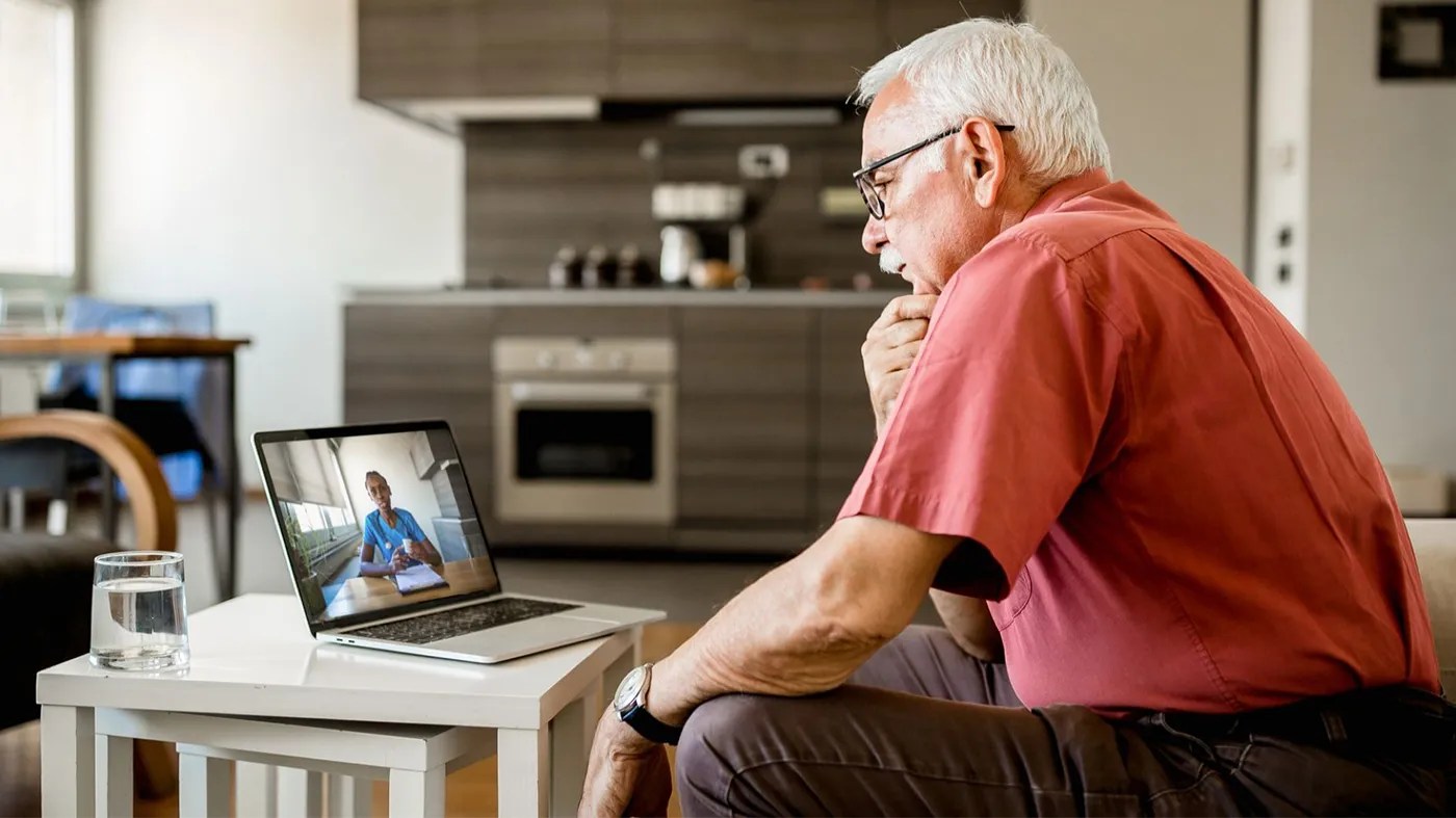 An elderly man in a red shirt attentively watches a video call with V A TelePulmonology on a laptop in a modern kitchen. A glass of water sits on a white table near him.
