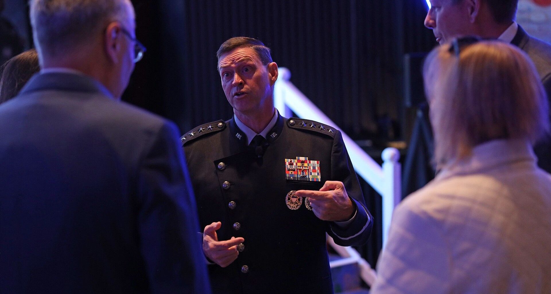 a man in a black military uniform speaks to a group of attendees on a conference ballroom floor