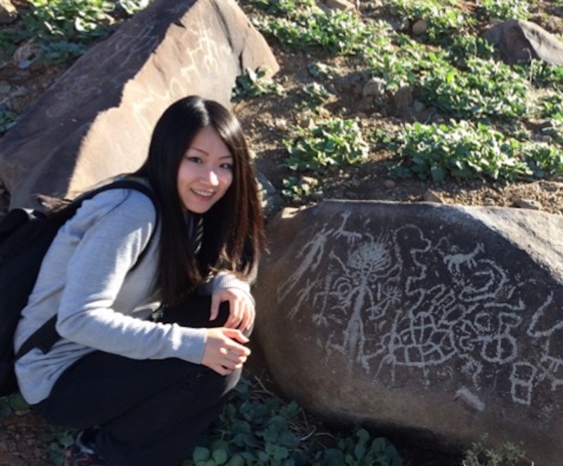 Smiling young woman with long, shiny black hair crouching beside a large rock with symbols marked on it.