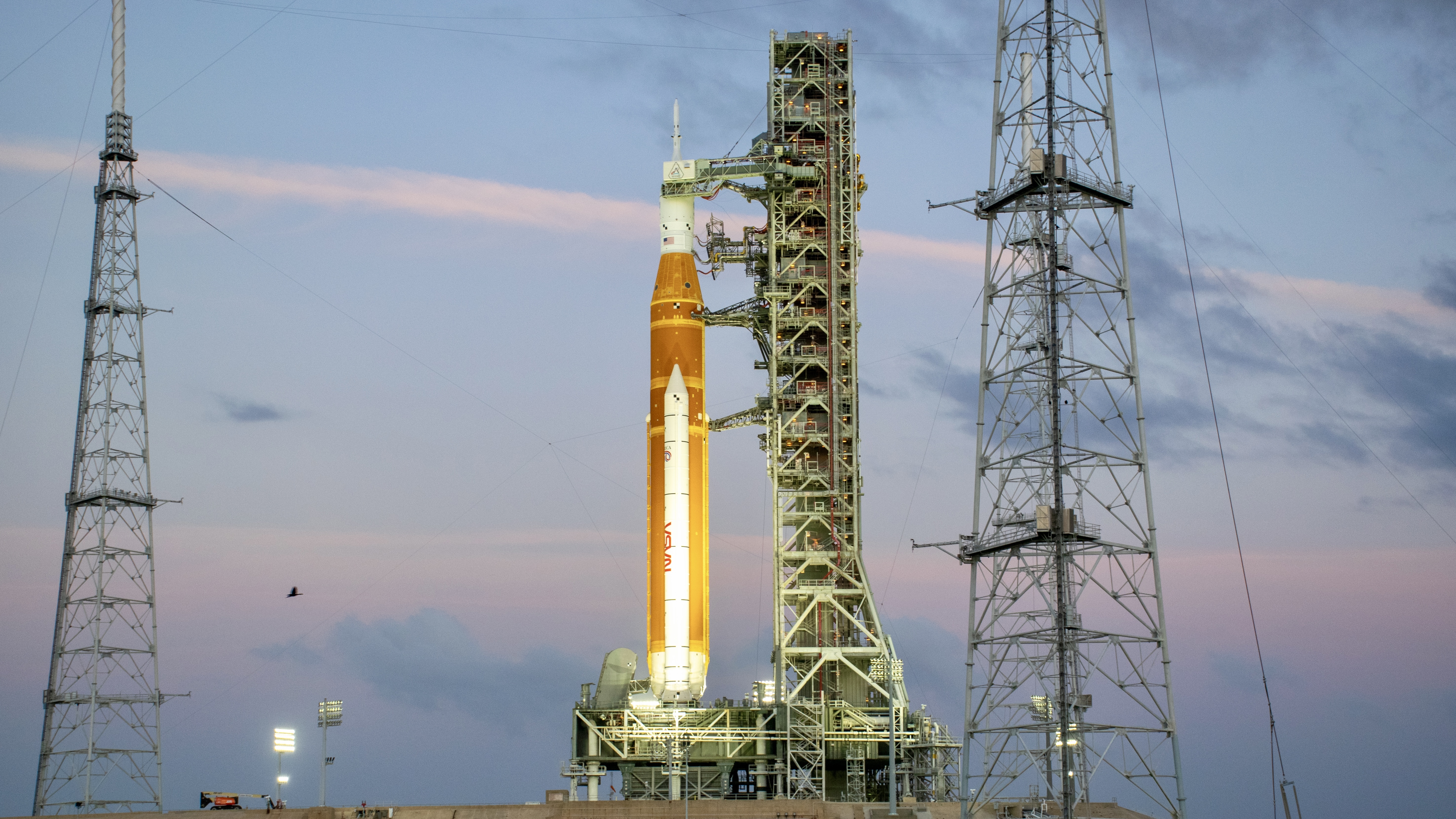 An orange rocket stands with a metal launch tower against a blue and pink sky.