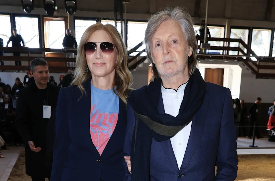 Two people on a runway. The woman wears sunglasses and a stylish suit with a graphic top. The man wears a classic suit with a scarf