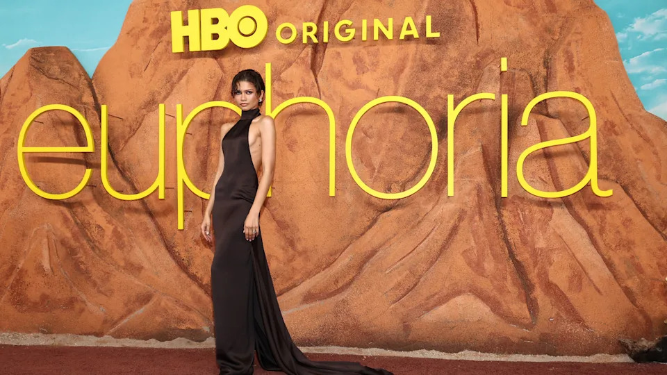 Zendaya poses in a sleek black dress at the "Euphoria" premiere, with large yellow text and a desert-themed backdrop.