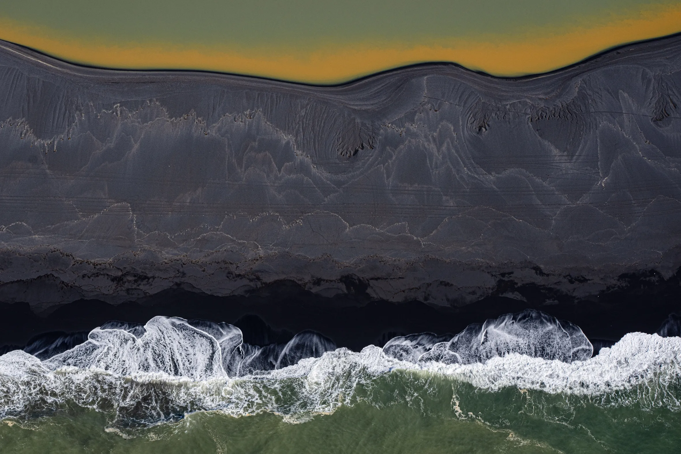 An aerial view of the southern coast of Iceland shows layered bands of basalt, black sand beach with patterns, and the ocean with white waves and green water.