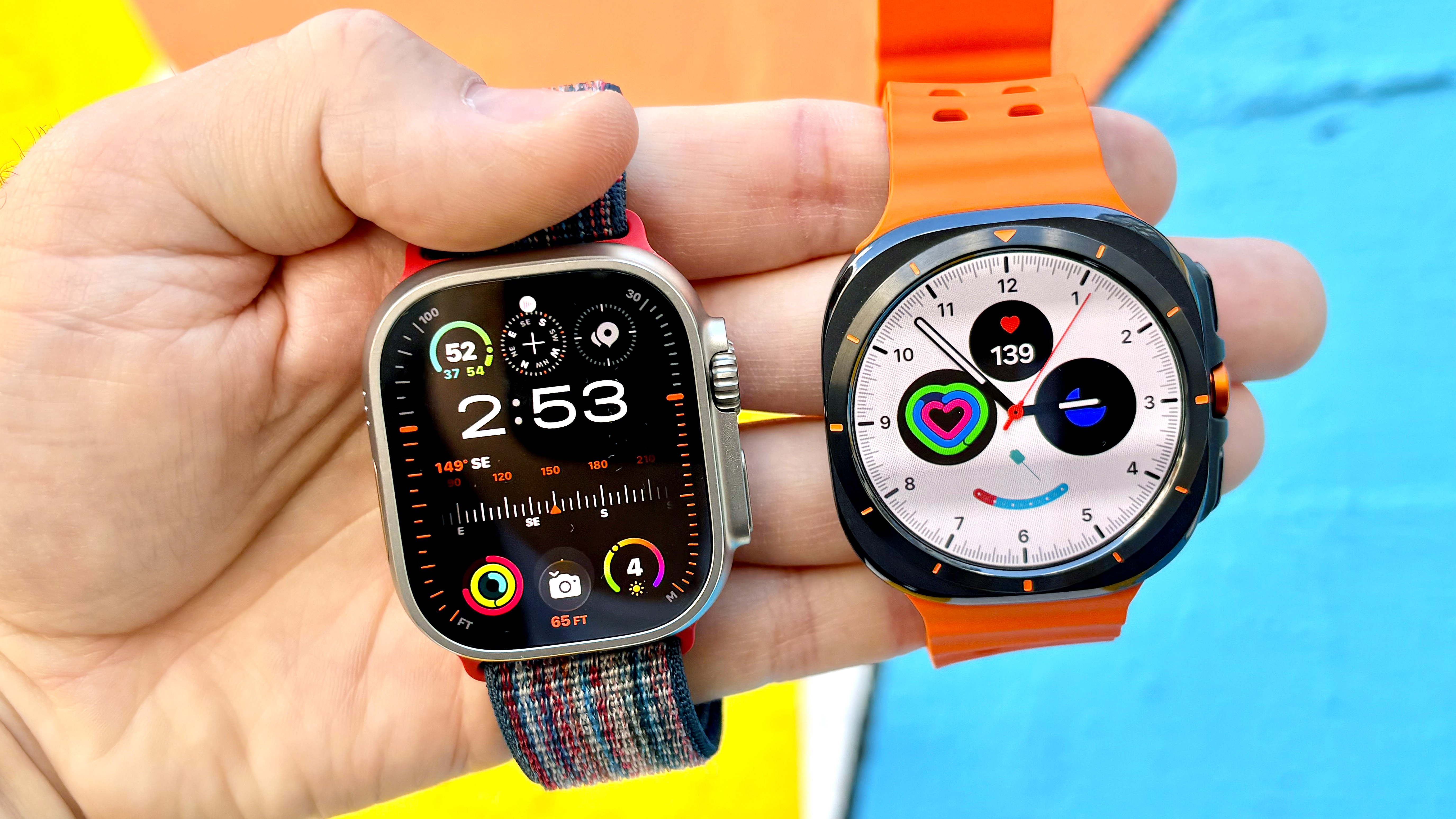 Close-up of the Apple Watch Ultra 3 next to the Samsung Galaxy Watch Ultra in the writer's hand against a colorful background