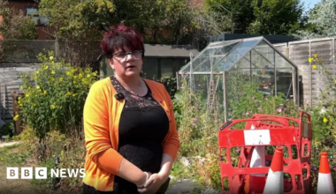 Melissa Stroud standing in front of a manhole with traffic cones and barriers around it. There are plants around a greenhouse behind it.