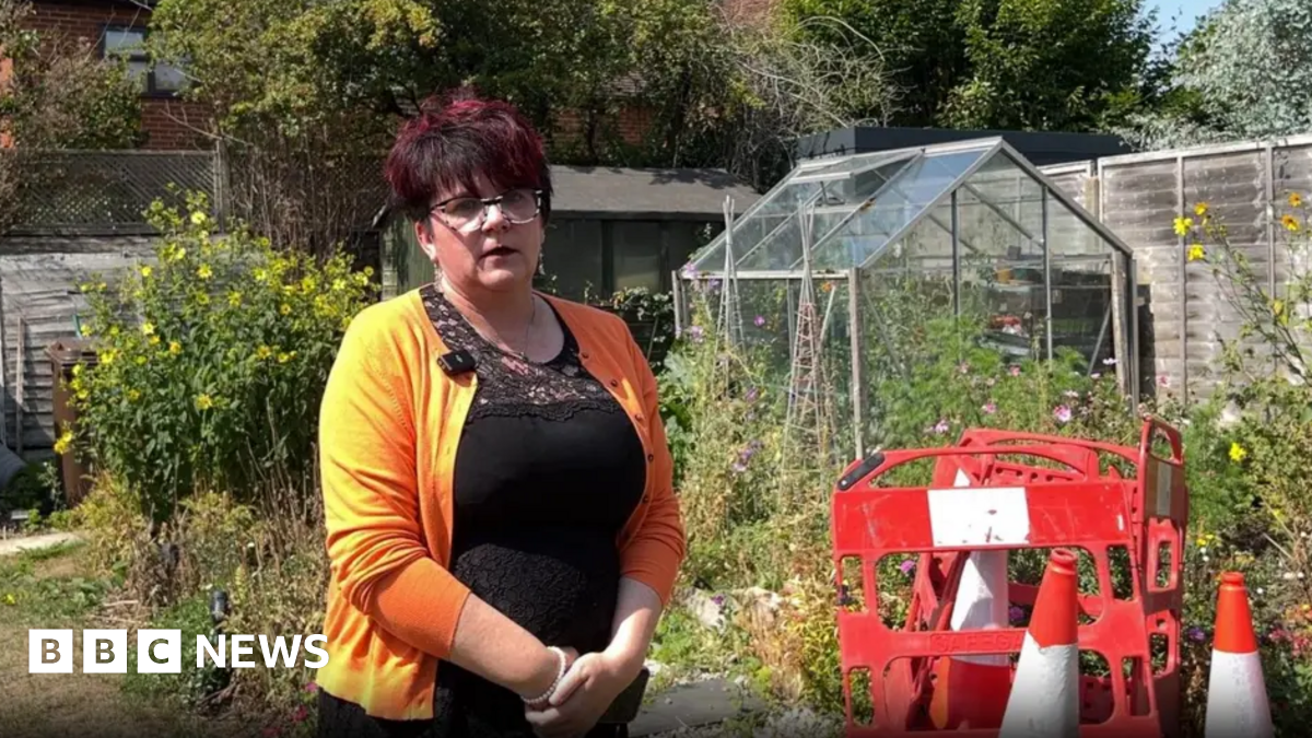 Melissa Stroud standing in front of a manhole with traffic cones and barriers around it. There are plants around a greenhouse behind it.