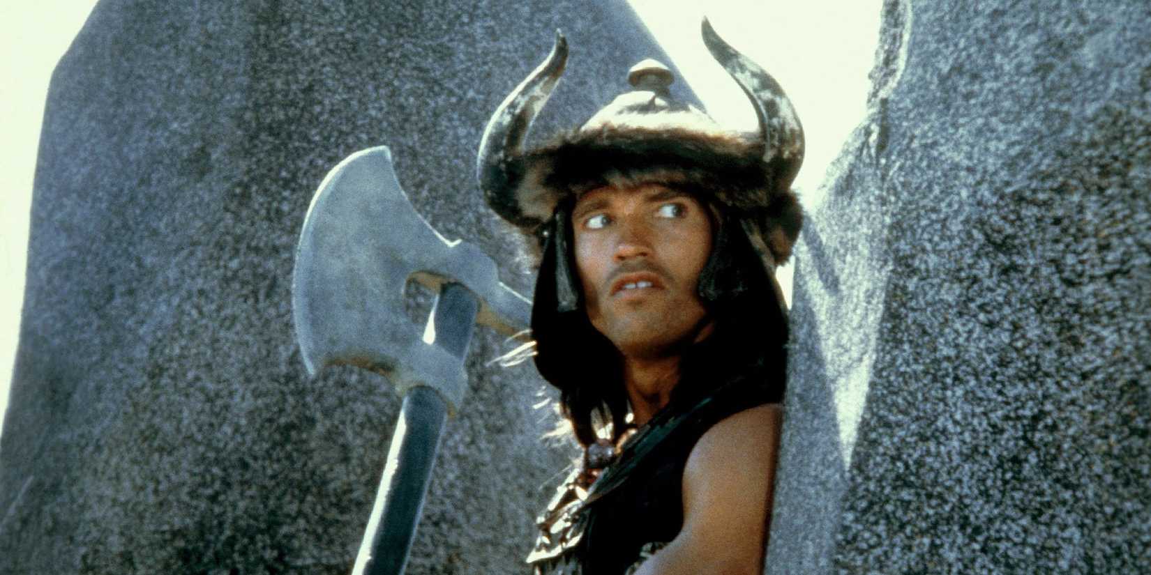 Arnold Schwarzenegger in Conan the Barbarian holding an axe and hiding between large rocks