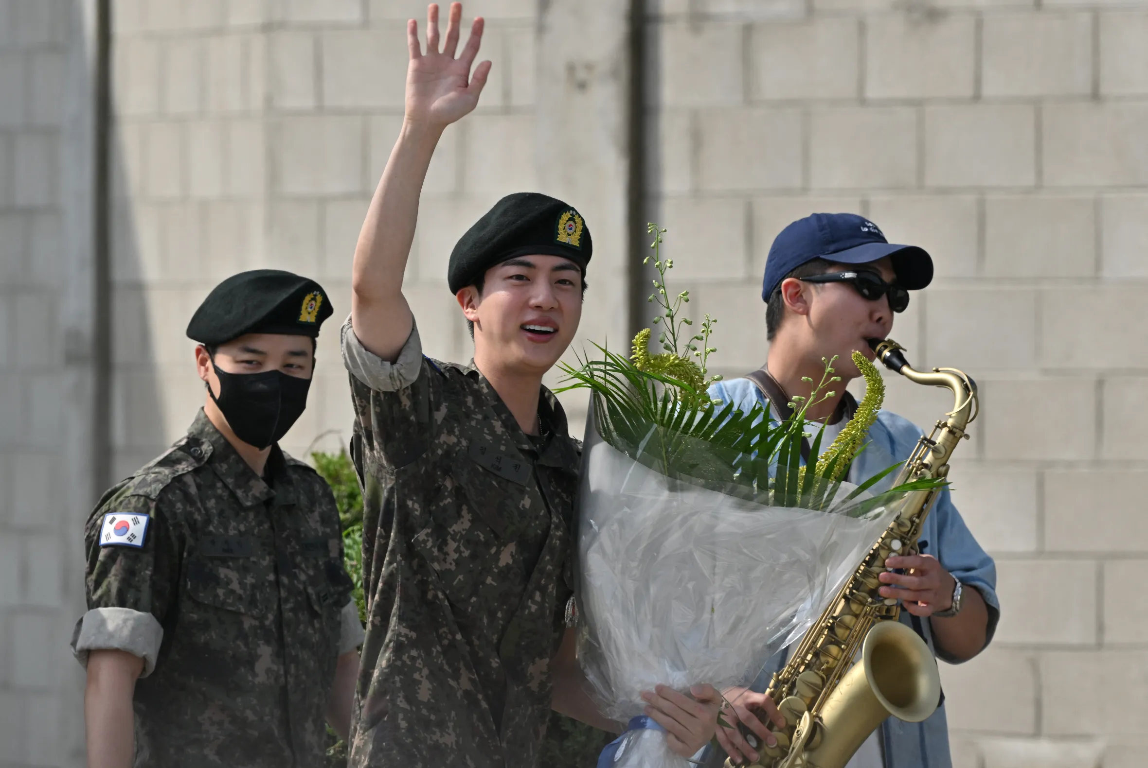 BTS member Jin in uniform waves, holding a bouquet, while fellow member RM plays the saxophone next to him.