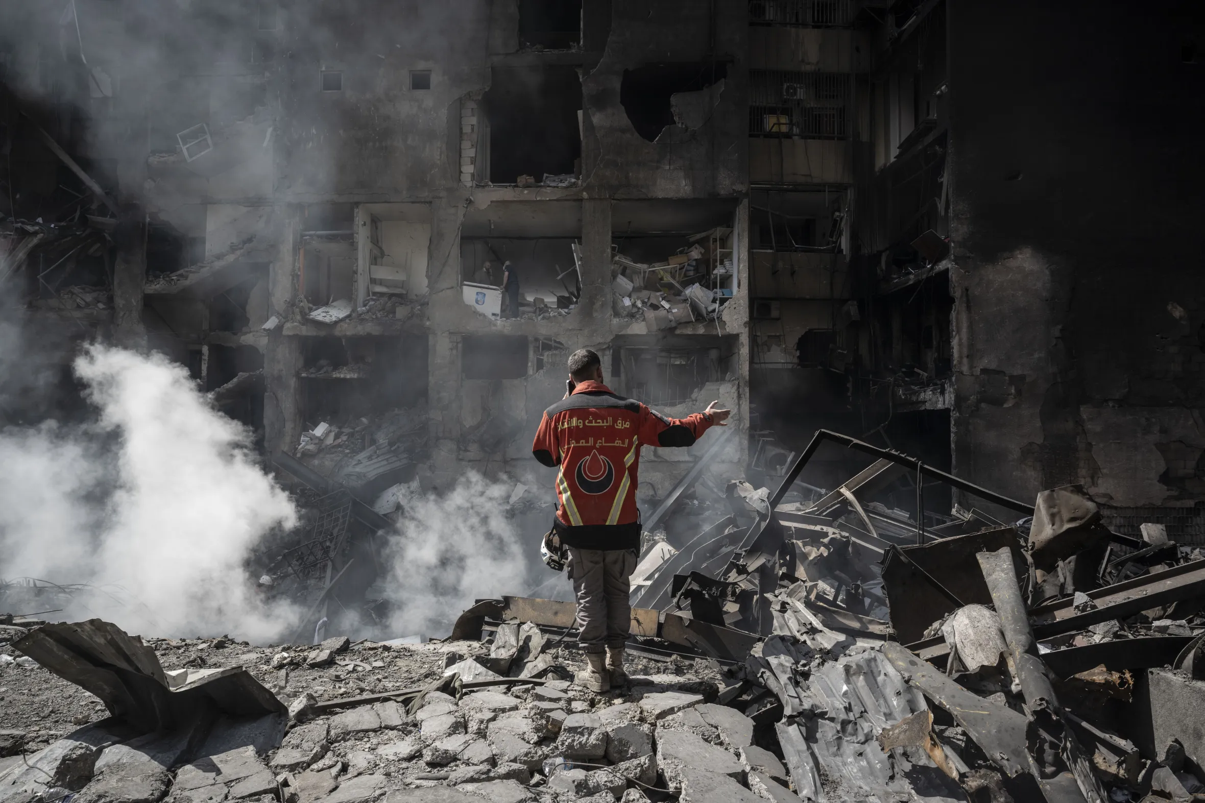 A first responder in an orange vest stands amid rubble and smoke from a damaged building in Beirut, Lebanon.