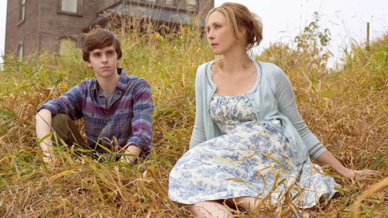 Norman Bates (Freddie Highmore) and Norma Bates (Vera Farmiga) sit on a grassy hill in front of their motel in Bates Motel