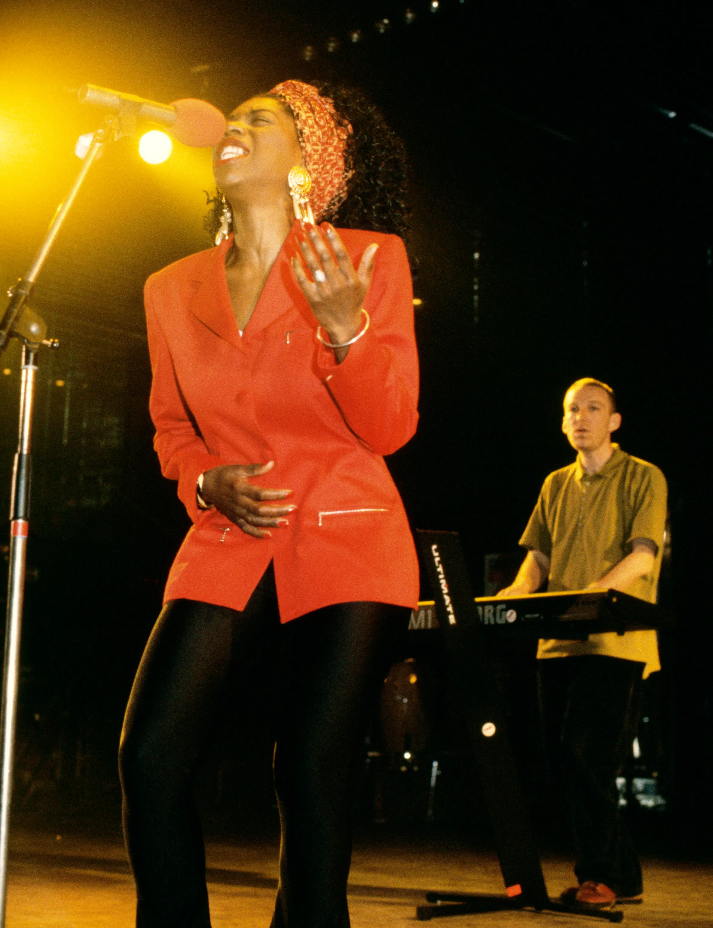 Heather Small singing into a microphone on stage, with Mike Pickering playing a keyboard behind her.