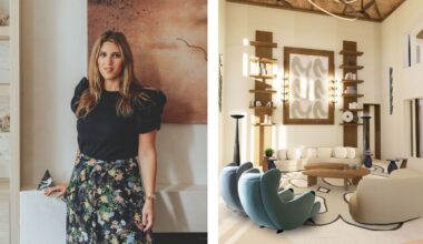 A high-ceilinged, contemporary living room designed by Lucy Goldbart featuring a neutral color palette with wood accents. Two curved cream sofas and two teal scalloped armchairs surround organic-shaped wooden coffee tables on a large patterned rug. The focal point is a large abstract wall hanging flanked by symmetrical wooden shelving units and tall, slender floor lamps.