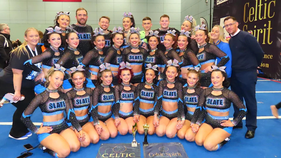 A number of cheerleaders and their coaches are standing side-by-side in a team photo. They are all dressed in their black and blue cheer uniforms with the word Slate written across their chests. The female members are all wearing ribbons in their hair. 
