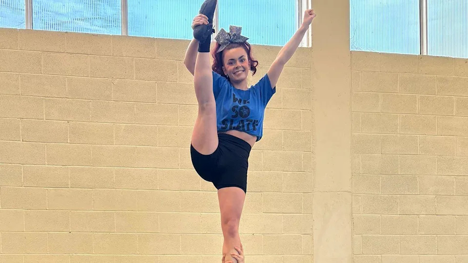 Clodagh McGonagle is standing holding her leg. She is wearing a blue top and black shorts. And has red hair tied up in a silver bow.