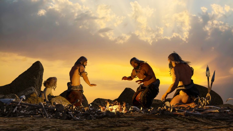 Realistic illustration of prehistoric people warming themselves by a campfire as the sun sets.