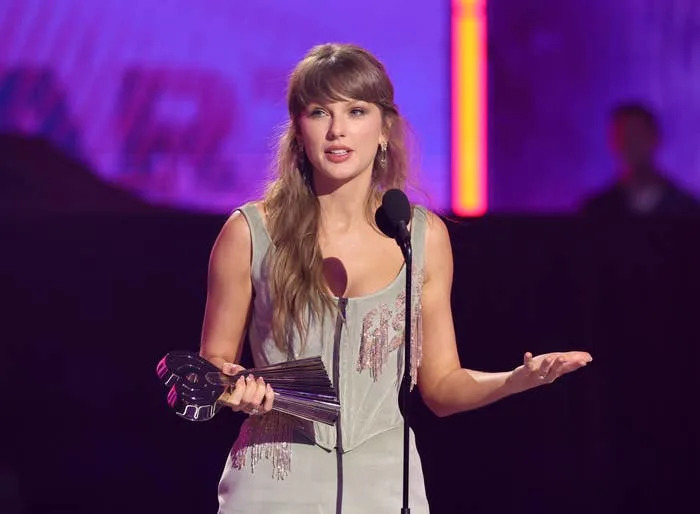 A celebrity in elegant attire holds an award while speaking at a microphone on stage