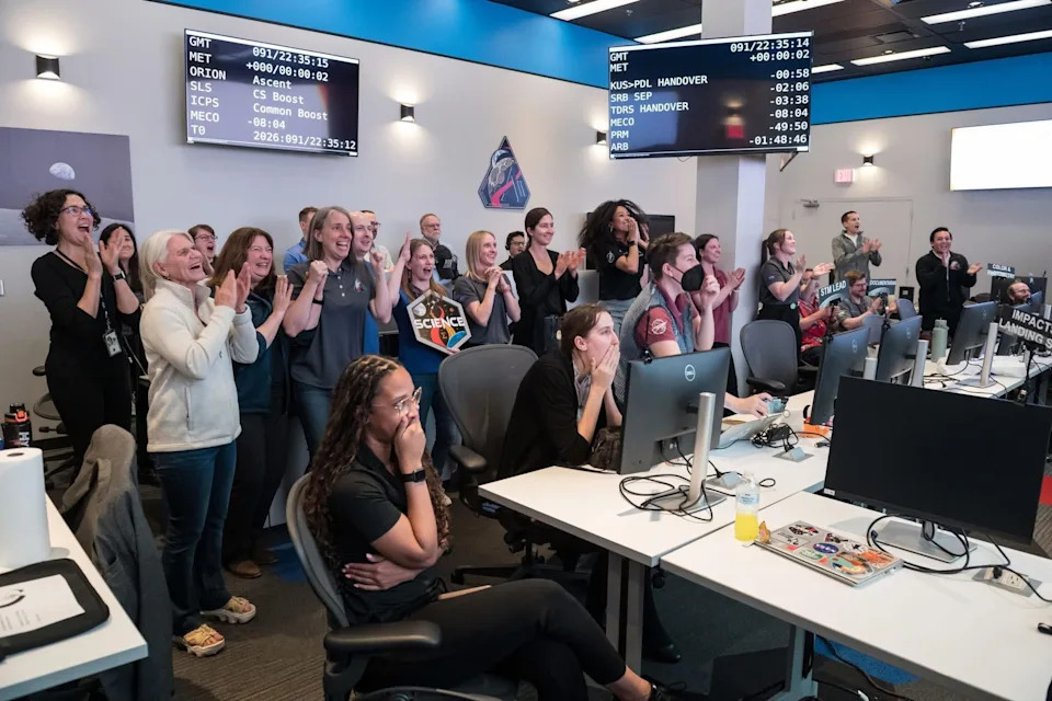 People in a control room cheer and applaud, watching a successful mission event on screens above. Various individuals display excitement and relief