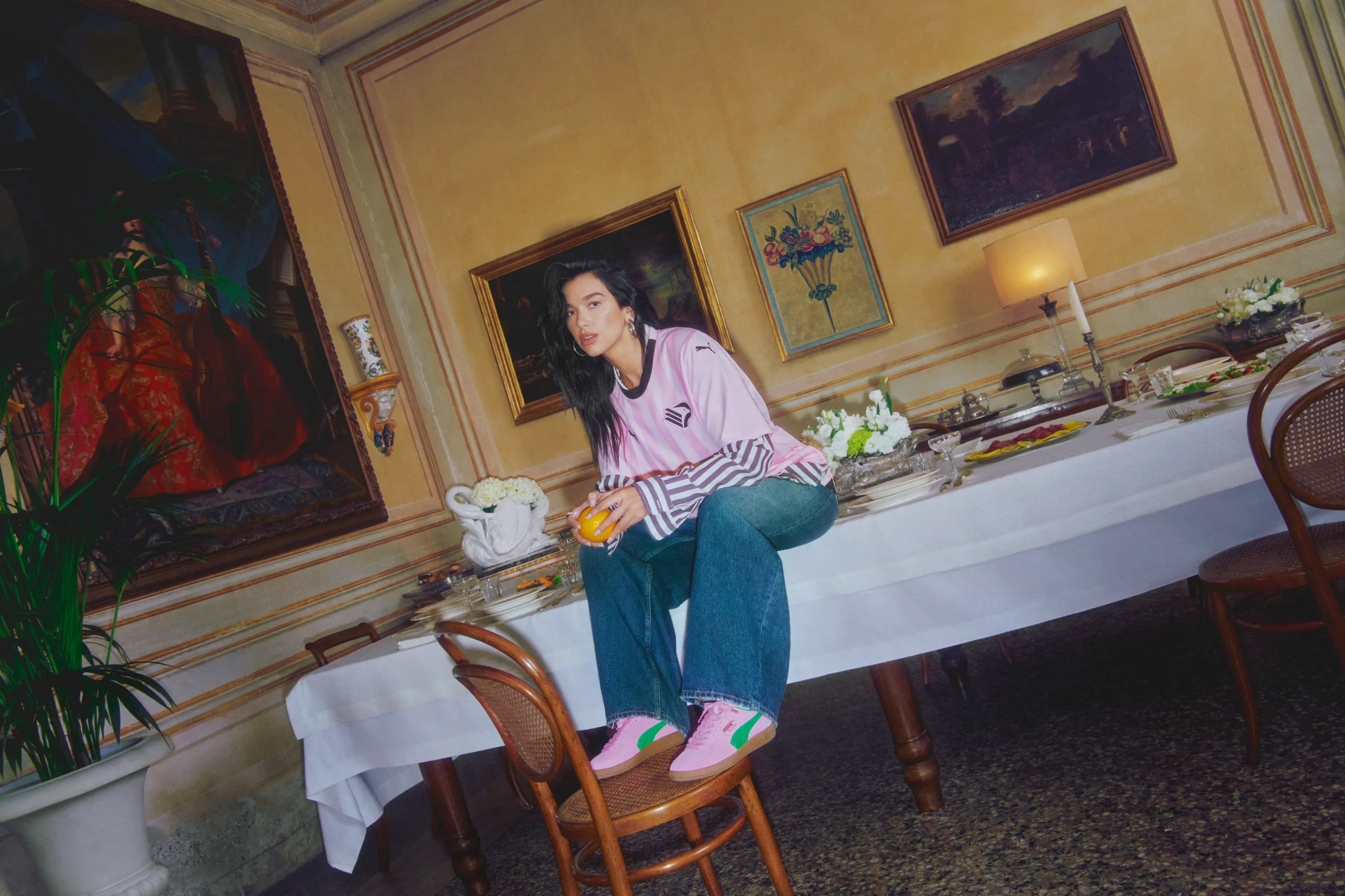 Dua Lipa sits on a long white dining table set with food, wearing a pink Puma athletic shirt, jeans, and pink Puma sneakers with green accents, holding an orange.