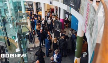 A crowd of people can be seen from above standing inside a building. There is a door to the left and columns through the middle. A large black sign is in front of the doors.