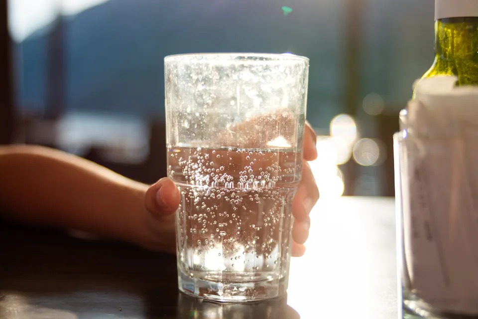 A hand holding a sparkling water glass with bubbles rising inside, sunlight glinting off the water, creating a refreshing and serene ambiance