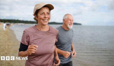 Active senior couple runners jogging outdoors on sandy beach by sea in early morning.