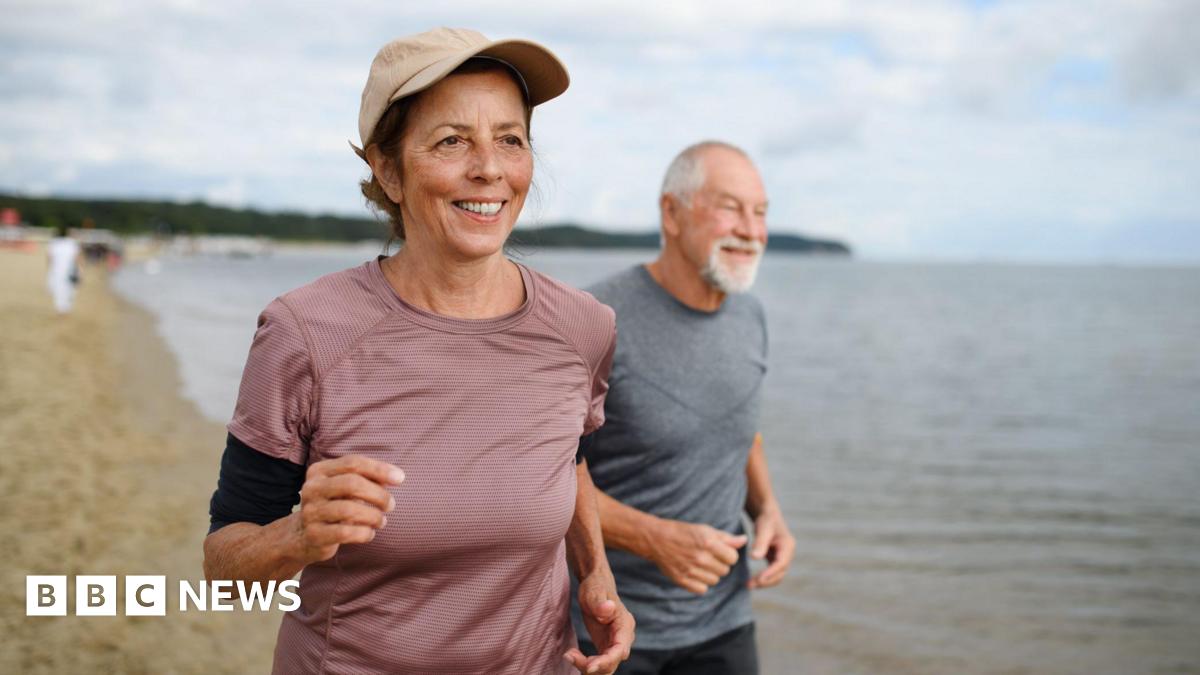 Active senior couple runners jogging outdoors on sandy beach by sea in early morning.