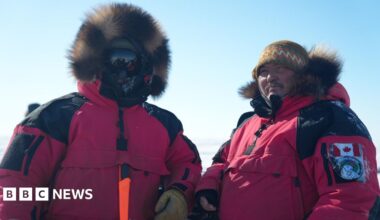 Two men -in the red, fur-trimmed parka uniforms of the Canadian Rangers. One has his face completly covered by shaded goggles, the other wears a hat and has his hood down.