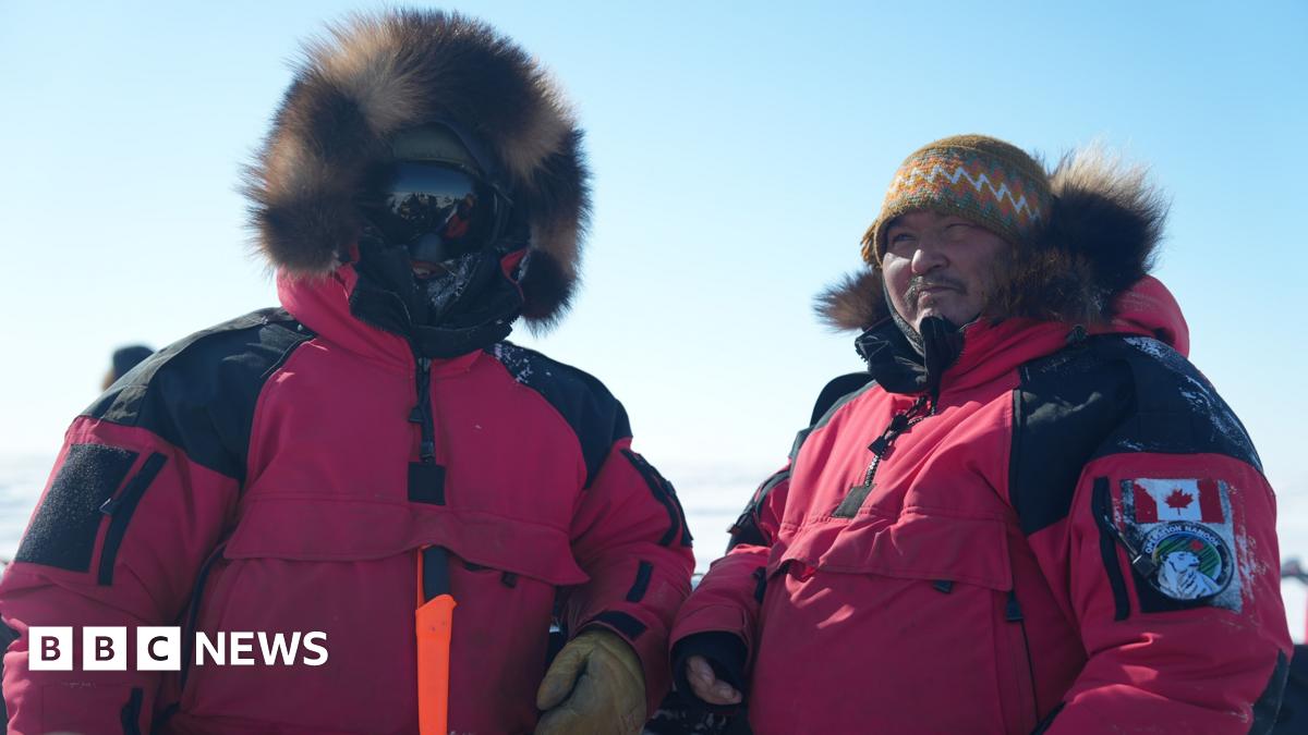 Two men -in the red, fur-trimmed parka uniforms of the Canadian Rangers. One has his face completly covered by shaded goggles, the other wears a hat and has his hood down.