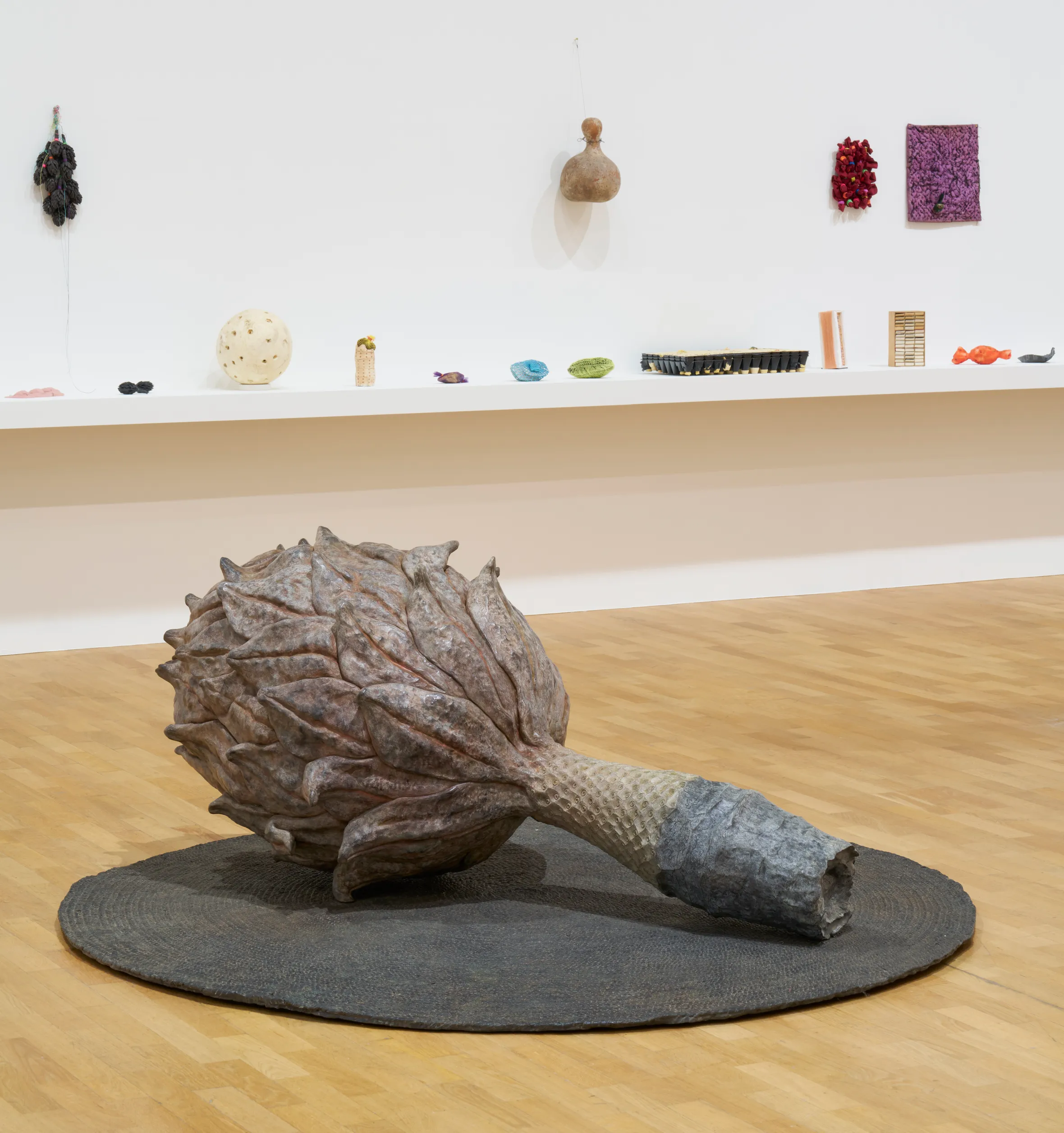 Installation view of “Veronica Ryan: Multiple Conversations,” featuring a large bronze magnolia pod sculpture on a round dark mat in the foreground and a shelf with various small sculptures against a white wall in the background.