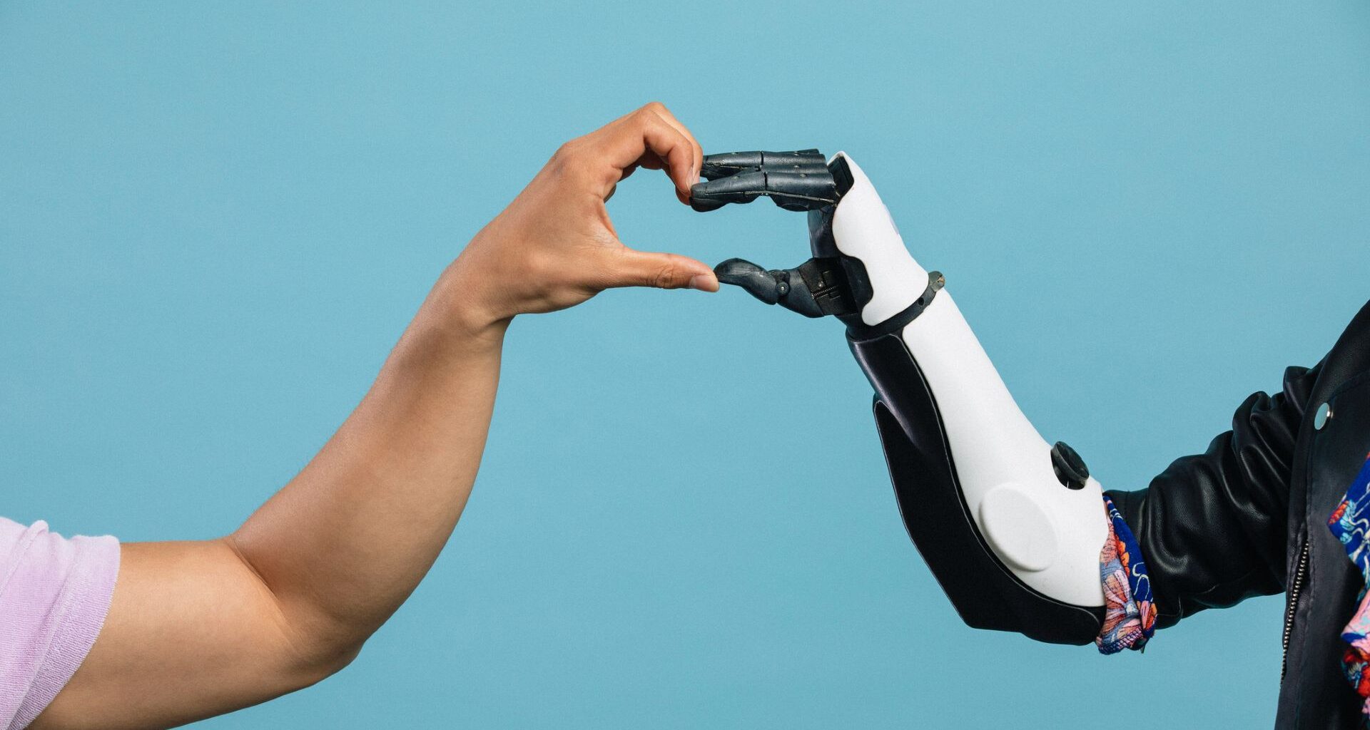 A person's left hand comes from left of the image to meet a black and white robotic hand from the right of the image to make a heart with their hands in the center, all in front of a blue background.