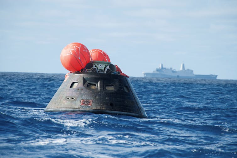 A spacecraft crew capsule floating in the ocean, with a large ship in the background.