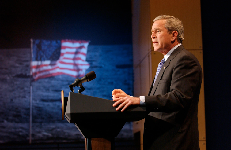 George W. Bush standing at a podium with an image of the US flag on the lunar surface in the background.