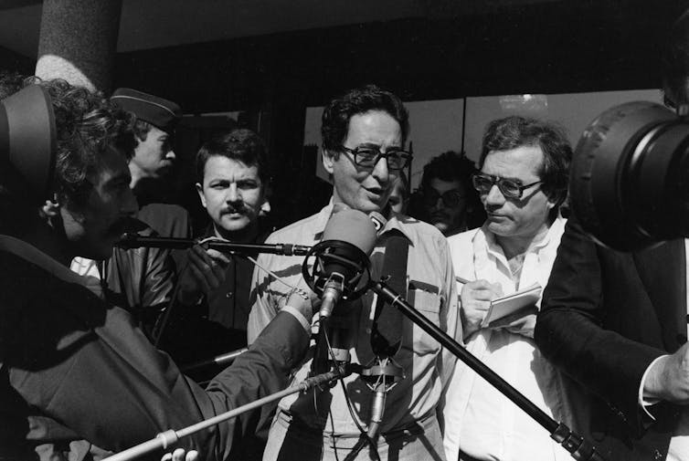 A man in glasses stands behind microphones