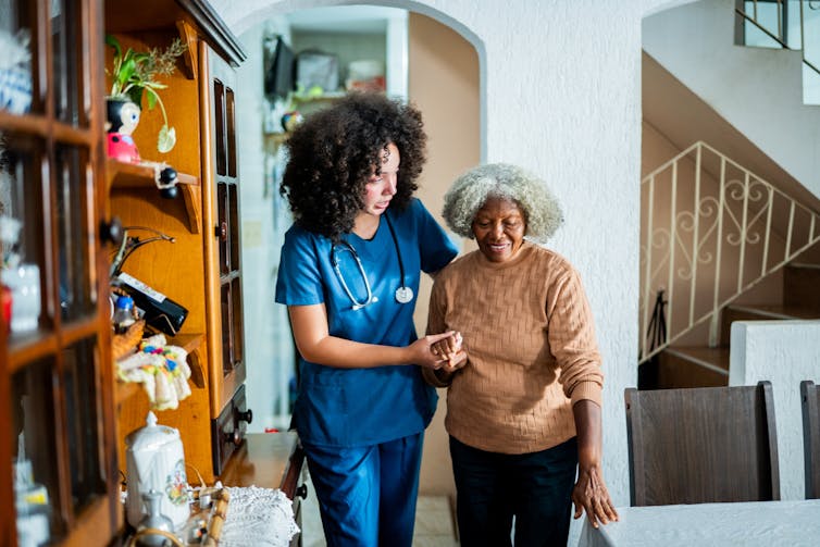 A home health aide helps a patient walk around a private home.
