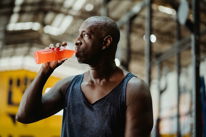 Healthcare worker drinking an orange sports drink during a workout in an indoor gym with industrial lighting.