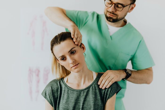 Healthcare worker in green scrubs examining a patient's neck and shoulder during a clinical assessment.