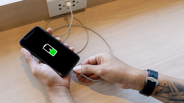 Hands holding a phone plugged into a charger with battery icon visible on the screen.