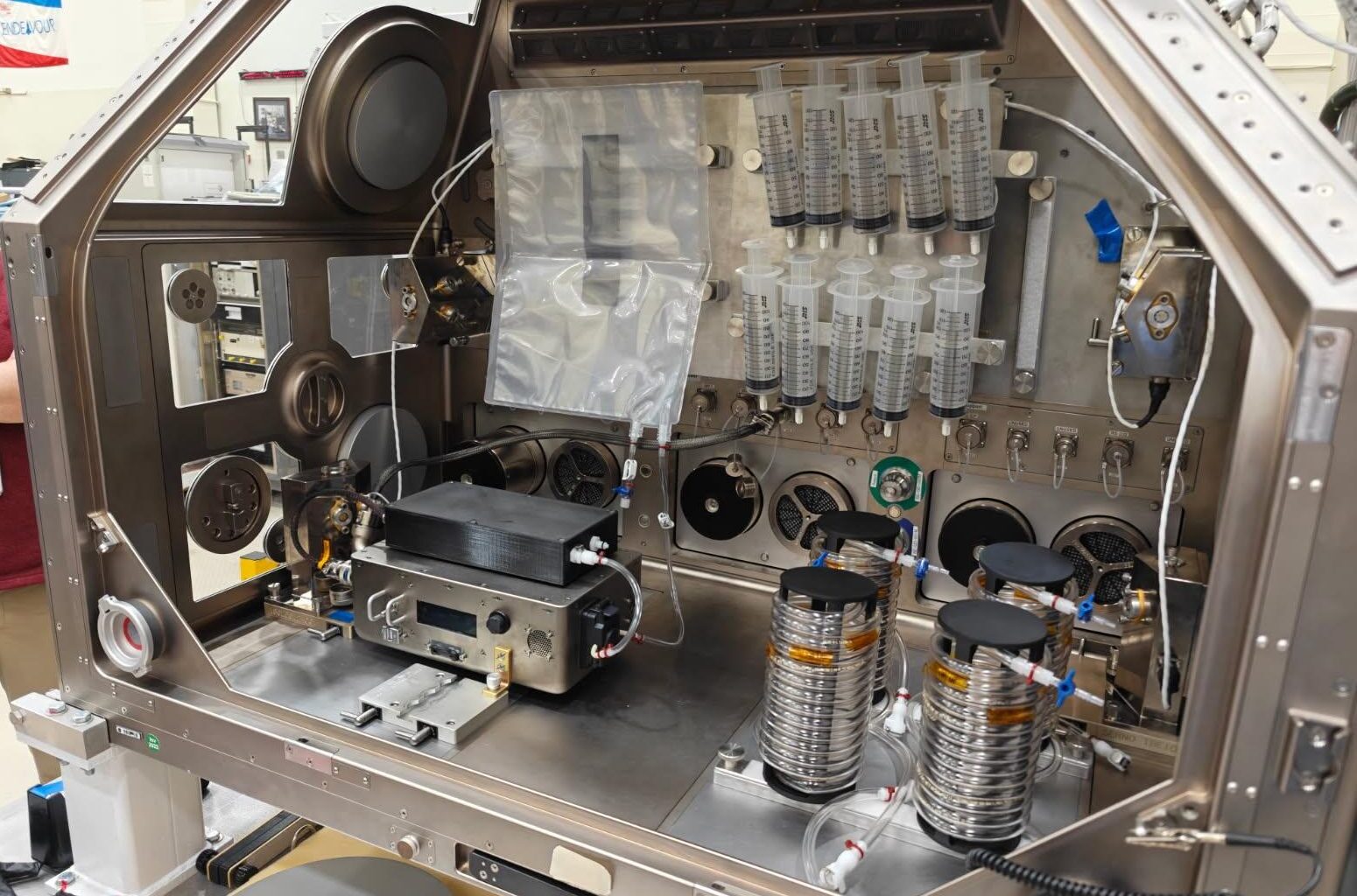 A large metal housing box containing the IVGEN Mini system is set up to create IV fluid. It includes the filtering system (two small boxes with cords attached), one plastic input bag, one plastic output bag, 10 syringes, additional cording, and four silver cylinder-shaped pieces of equipment.