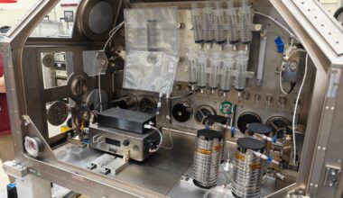 A large metal housing box containing the IVGEN Mini system is set up to create IV fluid. It includes the filtering system (two small boxes with cords attached), one plastic input bag, one plastic output bag, 10 syringes, additional cording, and four silver cylinder-shaped pieces of equipment.