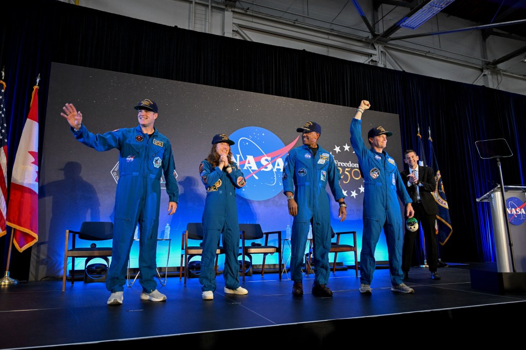 The four astronauts of Artemis II—each wearing blue flight suits and black baseball caps—stand on-stage, waving to the crowd at a welcome-home event.