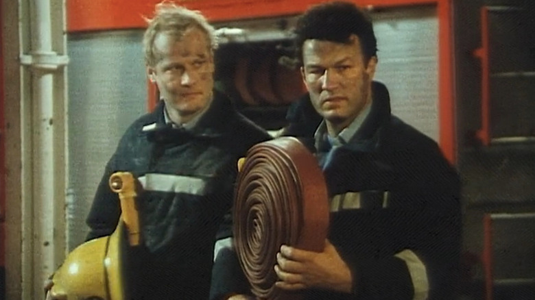 Rupert Baker as Malcolm next to Glen Murphy as George standing in firehouse with house and helmet in London's Burning